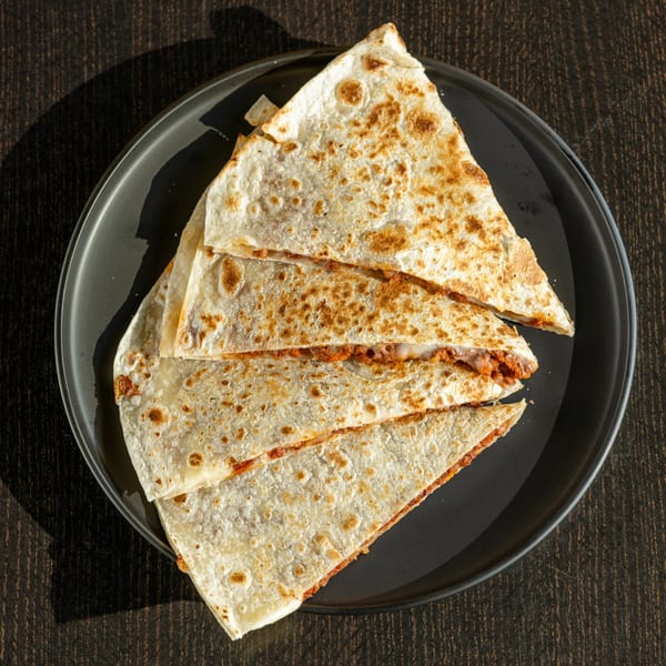 Four triangular slices of a golden-brown quesadilla filled with meat and cheese on a dark plate.