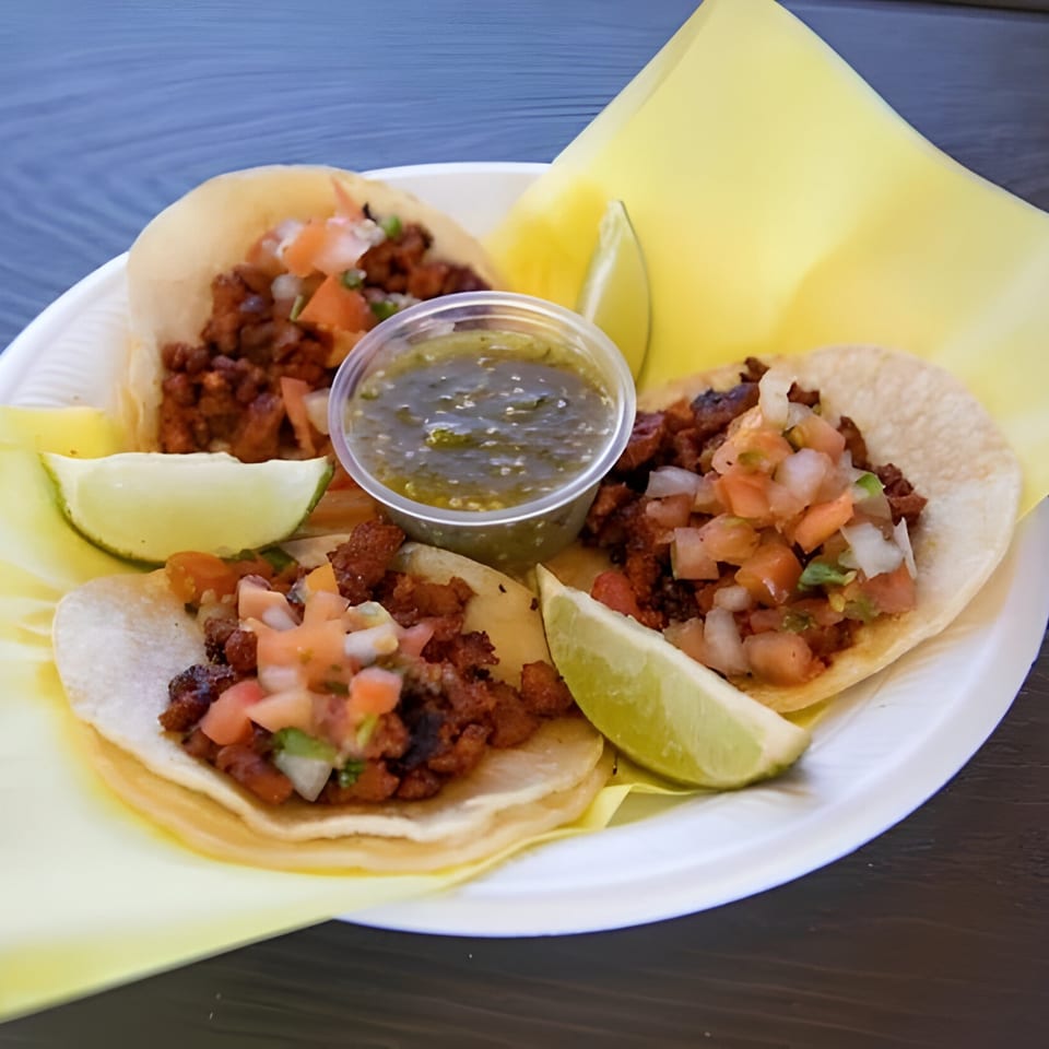 Three Mexican street tacos with meat, pico de gallo, lime wedges, and green salsa on a white plate.
