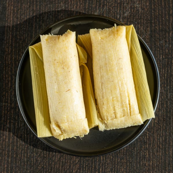 Two golden tamales, still in their corn husk wrappers, rest on a dark plate.